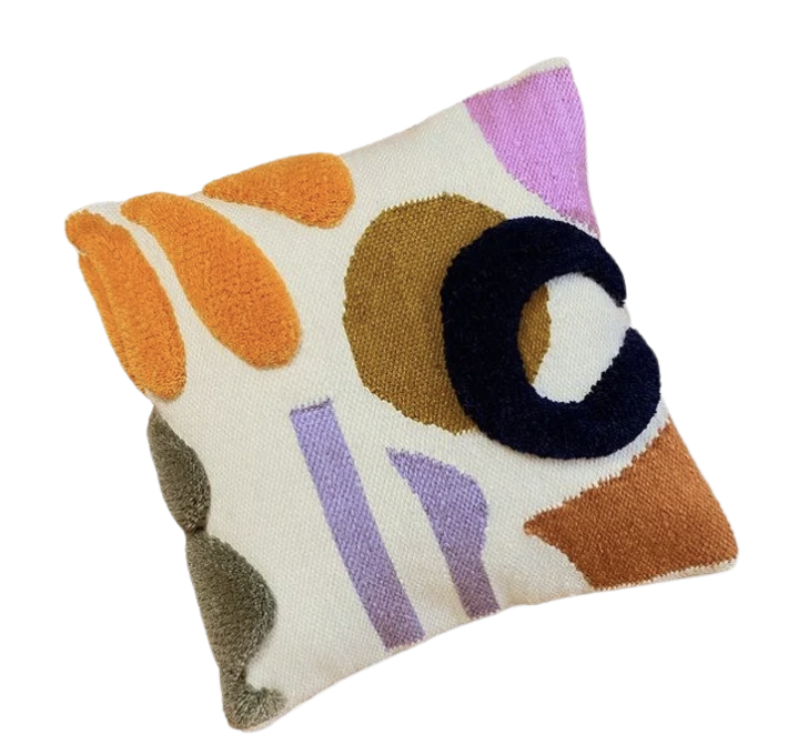 A close-up shot of a square throw pillow with a textured, abstract design. The pillow has an off-white background. Various shapes and colors are embroidered onto the pillow, some appearing raised. From top to bottom and left to right, the design elements include: In the top right corner, a block of solid light purple/pink. Below it, a large, dark brown or mustard-colored circle. Overlapping the right side of the circle are two thick, concentric black rings that appear to be made of a looped or fuzzy material. To the left of the brown circle, a cluster of three elongated, rounded shapes in bright orange, resembling petals or leaves. These also appear raised. Below the orange shapes, in the lower left quadrant, is a cluster of dark green or olive-colored rounded shapes, similar in texture to the orange ones. In the middle-left, between the orange and green shapes, are two vertical, somewhat irregular rectangular bars in a soft lavender color. In the bottom right corner, a block of burnt orange or rust color, partially visible. The overall impression is a modern, playful, and tactile design.
